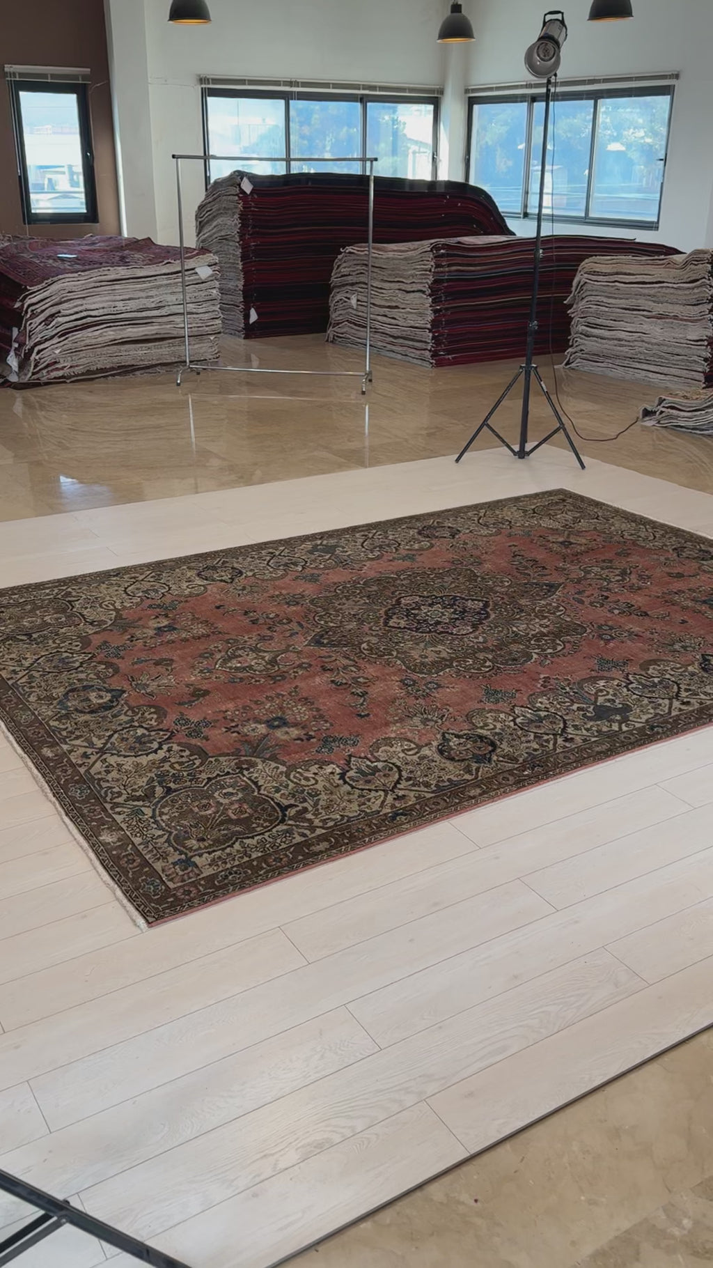 Vintage large wool area rug displayed in showroom with stacks of rugs in background, showcasing its rich faded red and brown hues.
